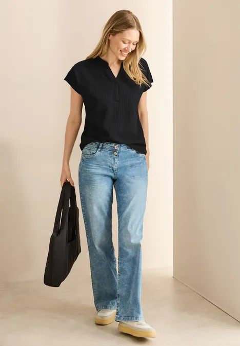 Frau mit schwarzem Shirt, Jeans und Tasche lächelt, steht in hellem Raum.